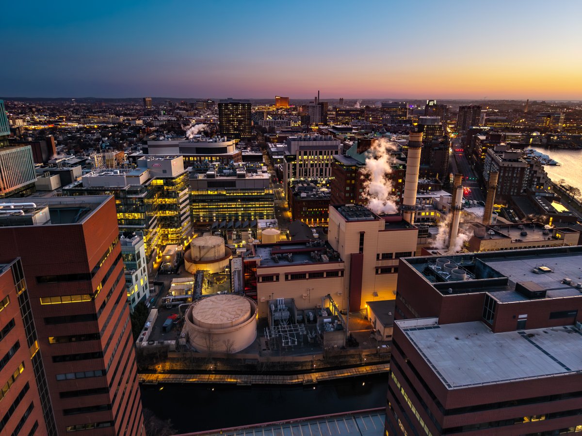 Aerial evening view of urban industrial infrastructure with power generation facilities, steam stacks, manufacturing buildings, and waterfront operations integrated into a modern city landscape.